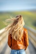 © songsi - Young woman with long hair wearing leather jacket on scenic pathway under blue sky with soft clouds and green hills