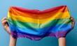 © Yuliia - Hands holding up colorful rainbow pride flag against blue background, symbolizing LGBTQ+ community support, equality, and celebration of diversity.