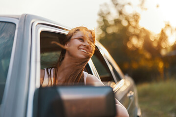 Wall Mural - Young woman enjoying a summer road trip, smiling joyfully with wind in her hair while looking out of the car window in a lush green landscape.