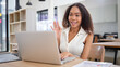 © wattana - Smiling young woman waving during an online video call on her laptop, sitting at a modern workspace.