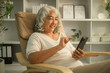 © wattana - Smiling senior woman engaging with her smartphone while relaxing in a modern rocking chair.