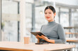 © kenchiro168 - Asian woman sitting at table in modern cafe, using tablet and smiling.