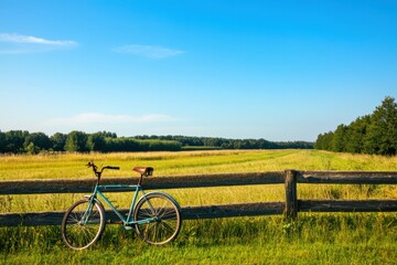 Poster - Bicycle rests against wooden fence by open field under bright blue sky during afternoon light. Generative AI