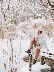 Beautiful young girl on a bridge near a lake and drinking hot tea in winter forest