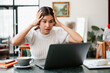 © Satori Studio - A young woman looks stressed while working on her laptop at home, surrounded by a cozy and modern living room setting.
