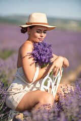 Wall Mural - woman sitting in a field of lavender and wearing a straw hat. She is smiling and holding a bouquet of flowers. Scene is peaceful and serene, as the woman is surrounded by the beauty of nature