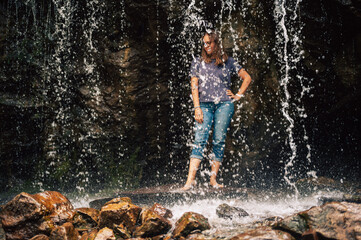 A woman in casual clothes stands barefoot behind a cascading waterfall, surrounded by rocky terrain, enjoying nature and the refreshing water.