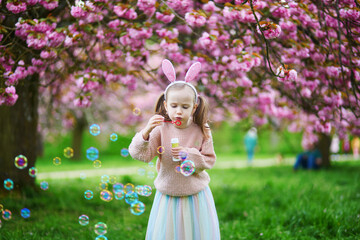 Wall Mural - Adorable preschooler girl wearing bunny ears blowing bubbles and enjoying nice spring day in cherry blossom garden.