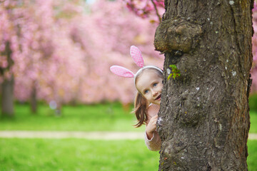 Wall Mural - Adorable preschooler girl wearing bunny ears enjoying nice spring day in cherry blossom garden.