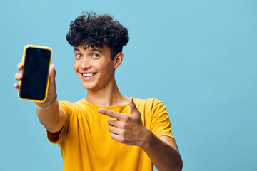 Wall Mural - young man showing smartphone, cheerful expression, bright yellow shirt, blue background, modern lifestyle, technology, social media engagement, casual fashion, youth happiness