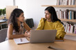 © fizkes - Peer mentorship. Smart young girl college university student support help female groupmate in learning research teach to use education app on notebook pc at library share idea at self preparation time