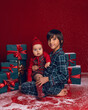 © saulich84 - brother and sister are sitting on the floor among green New Year gift boxes on a red background with snow.