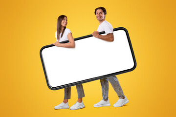 Wall Mural - Cool Mobile App. Cheerful young couple holding big giant smartphone with mockup, walking together, promoting application or website, advertising your service or product over orange studio background