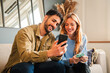 © Jose Calsina - Happy couple enjoying time together, sharing a moment while looking at their smartphones with smiles and laughter in a cozy home living room, capturing the joy of relationships and technology