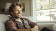 © VALUEINVESTOR - A happy man sitting on a couch at home, looking to the side and smiling in living room with a window, generative AI
