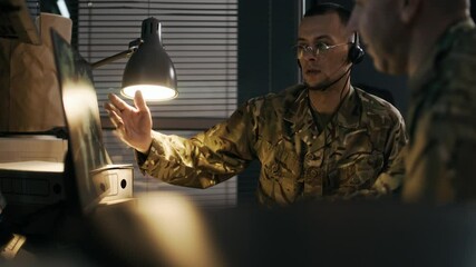 Handheld shot of two cybersecurity military agents in camouflage uniform sitting in front of computer screen while observing program with global map in command center