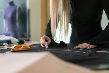 A textile artist carefully uses chalk to draw lines on black fabric, surrounded by various sewing tools and materials in a well-lit studio.