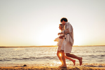 Wall Mural - Smiling beautiful woman and her handsome boyfriend. Excited couple in casual clothes. Happy cheerful family. Female and man run at sunrise over sea beach outdoors. Run at seaside in summer day