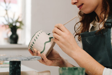 Poster - Female artist is painting earthenware with paintbrush and water colors in the workshop. Painter is drawing and making a colorful decoration of a clay ware.