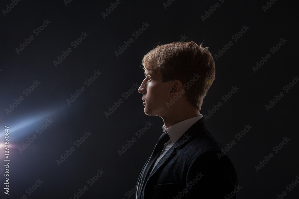 In a dark studio, a man in a black suit and white shirt stands in ...