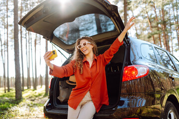 Wall Mural - Young woman is resting in trunk of car and taking selfie. Blogging, communication, technology. Travel by car