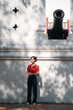 © Jirawatfoto - Asian woman in red shirt and dark pants stands in front of historic cannon, with dappled sunlight creating shadows on white wall behind her