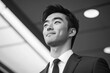 © Mane - Handsome Asian businessman in a suit, standing with a confident smile and looking away, against office background.