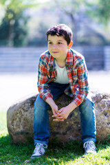 Casual boy sitting on a rock in a park with a thoughtful look.