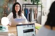 © Studio Marmellata - A young woman with curly hair wearing a white sweater is smiling while working on her laptop in a stylish workspace with clothing racks and decorations in the background.