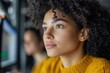 © Kate - Young woman with curly hair looks thoughtfully at her computer, immersed in creative work with a colleague nearby
