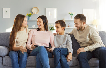 Happy young parents with two children boy and girl sitting on sofa using modern tablet together browsing internet and talking enjoying weekend. Family leisure and technology concept.