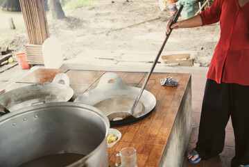 Southern Vietnamese lady in traditional dress stirring mixture of squeezed coconut milk, cream, sugar, malt syrup in flat cast iron pans at local handmade candy factory in Can Tho, control flame