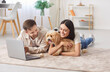 © Studio Romantic - Married young couple enjoying leisure time at home, using a laptop for online activities while playing with dog, lying on floor. The domestic happy entertainment and companionship with animal.