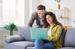 © Studio Romantic - Young family couple sitting at home, sofa looking smiling at laptop screen, happy young married spouse web surfing, making purchases online booking flight tickets, reading good news, leisure time