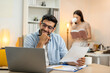 © Montri - Young businessman working at home office on laptop computer, finishing his online work, while his girlfriend and roommate sitting behind on sofa relaxing and waiting. Freelance business. couple.
