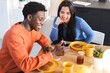 © Studio Marmellata - A man in an orange sweater and a woman in a blue sweater smile while sharing a breakfast of pancakes and coffee at a cozy dining table, engaging in a cheerful conversation.