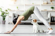 © Prostock-studio - Athletic asian young woman doing yoga with her adorable loyal red and white doggy at home, chinese lady standing in downward dog pose, jack russel puppy sitting by owner on fitness mat, side view