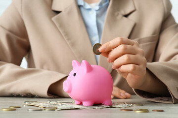 Wall Mural - Businesswoman putting coin into piggy bank at table, closeup. Financial Wellness Month