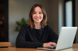 © He - Middle aged woman with laptop at wooden table smiles camera