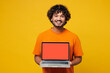 © ViDi Studio - Young smiling happy IT Indian man wear orange t-shirt casual clothes hold use work on laptop pc computer with blank screen workspace area isolated on plain yellow background studio. Lifestyle concept.