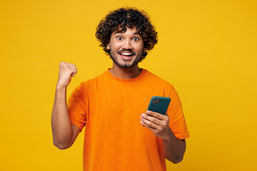 Young smiling happy fun Indian man he wears orange t-shirt casual clothes hold in hand use mobile cell phone do winner gesture isolated on plain yellow background studio portrait. Lifestyle concept.