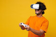 © ViDi Studio - Young gambling Indian man he wear orange t-shirt casual clothes hold in hand play pc game with joystick console watching in vr headset pc gadget isolated on plain yellow background. Lifestyle concept.