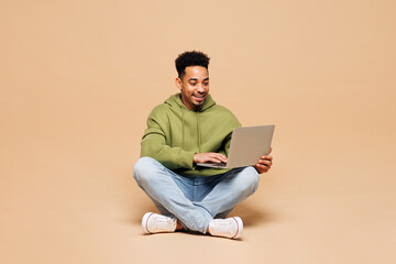Full body young smart IT man of African American ethnicity wears green hoodie casual clothes sit hold use work on laptop pc computer isolated on plain pastel light beige background. Lifestyle concept.