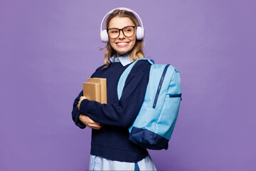 Poster - Side view young girl student wear blue casual clothes glasses backpack bag hold books listen music in headphones look aside isolated on plain purple background. High school university college concept.