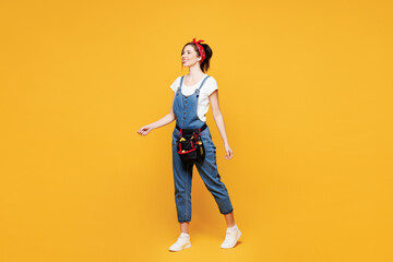 Poster - Full body side view young employee laborer handyman woman wears overalls belt walk go isolated on plain yellow background. Instruments accessories for renovation apartment room. Repair home concept.