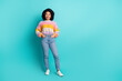 © deagreez - A stylish young woman in a colorful sweater and jeans stands confidently against a turquoise background