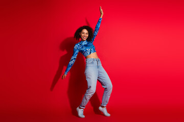 Wall Mural - Young fashionable woman joyfully posing against a vibrant red background wearing a stylish printed top and jeans