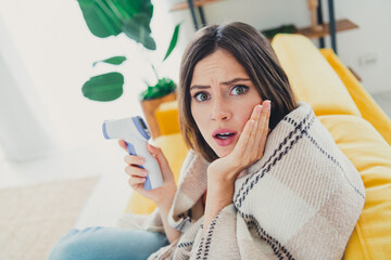 Wall Mural - Surprised young woman wrapped in blanket holding digital thermometer in living room