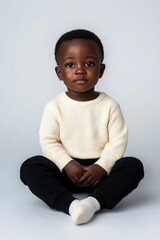 Wall Mural - A young black boy is sitting on the floor wearing a white sweater and black pants. He has a serious expression on his face