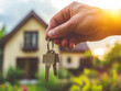 © Hlen - Close-up of a hand holding a house key, with a large house in the background. Associations with buying a house, real estate transactions and transfer of ownership